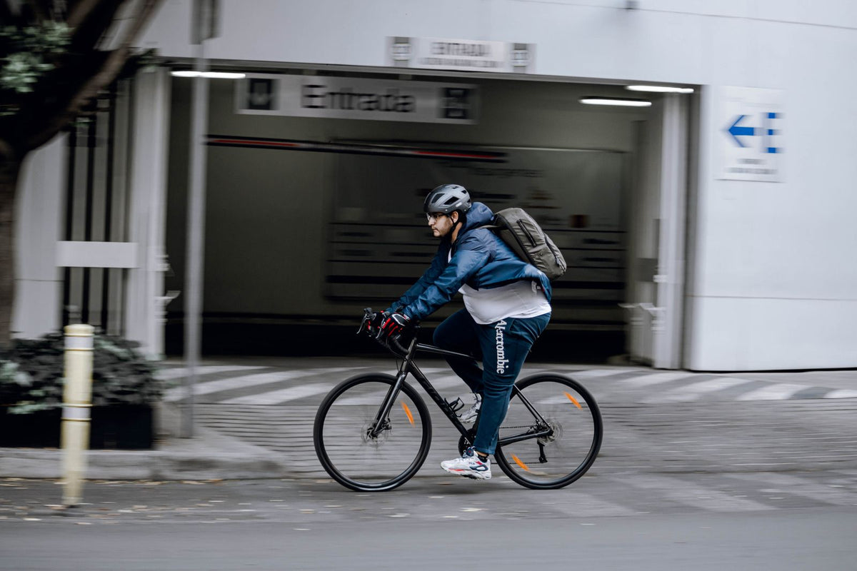 Un cycliste casqué, un sac à dos sur les épaules, roule dans une rue de ville devant l'entrée d'un parking.