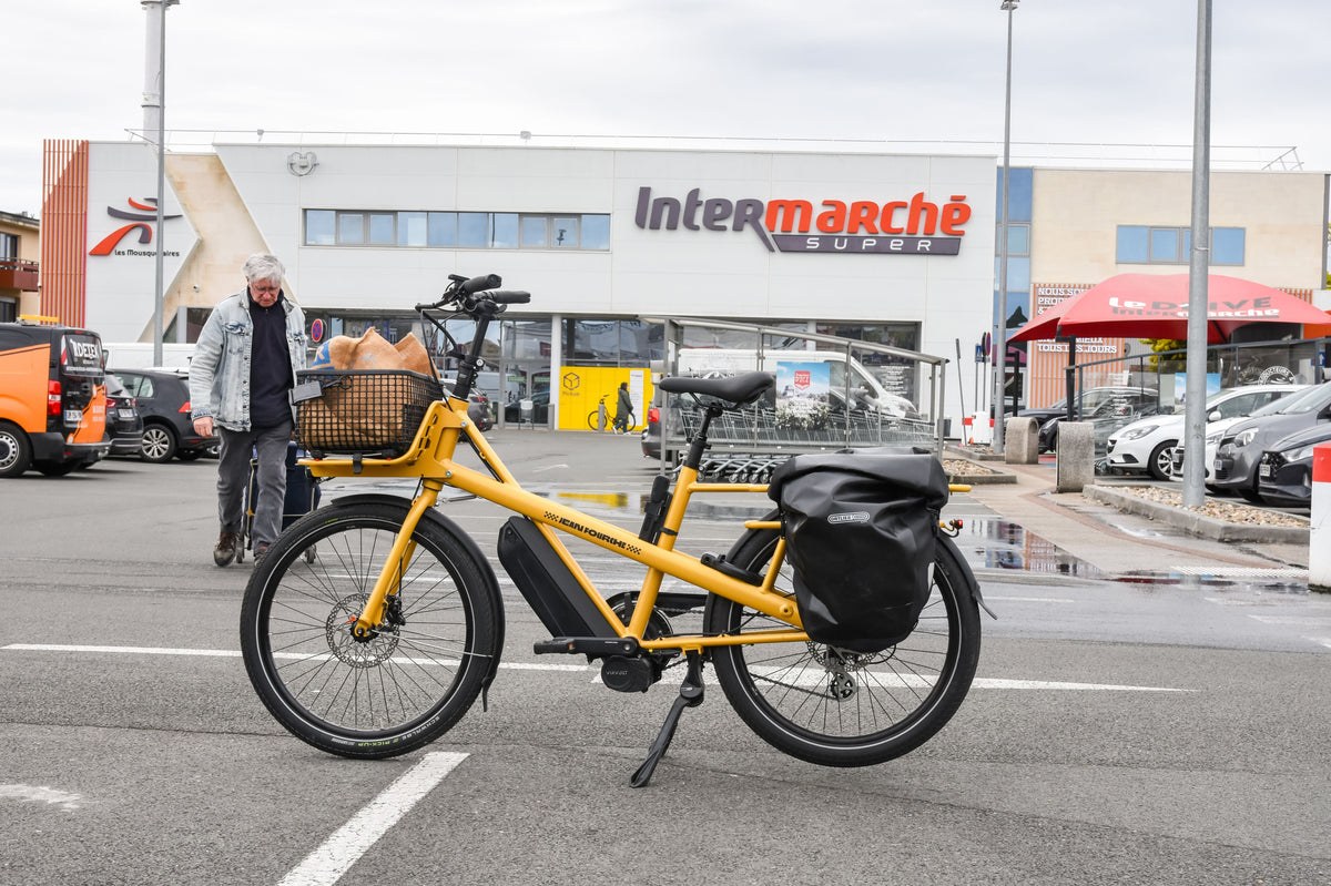 Vélo cargo électrique jaune stationné devant un supermarché, prêt pour le chargement des courses