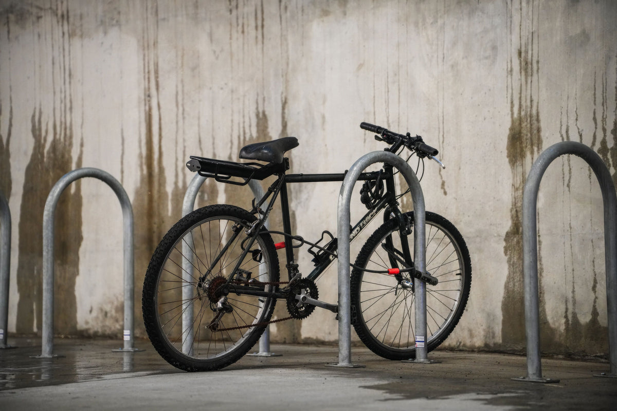 Vélo noir attaché par un antivol à un arceau de stationnement devant un mur de béton mouillé