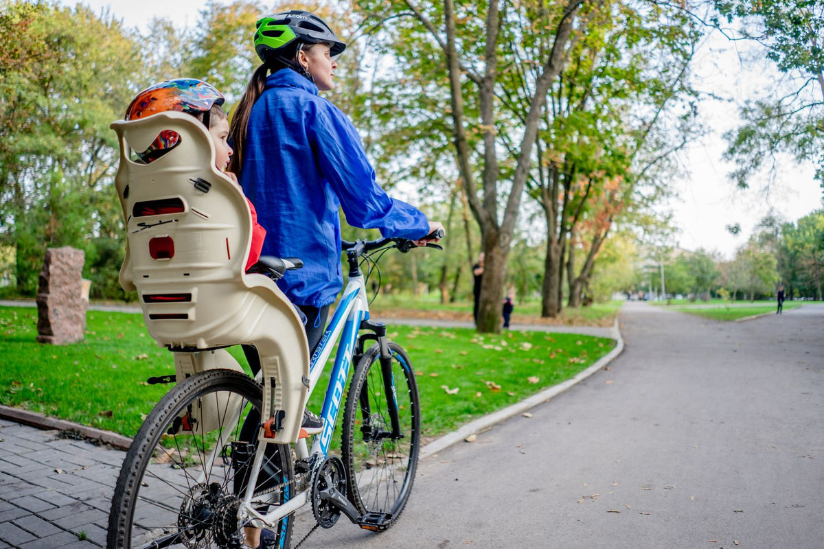 Adulte casqué à l'arrêt sur une allée de parc, avec un jeune enfant casqué installé dans un siège enfant à coque haute fixé au-dessus de la roue arrière